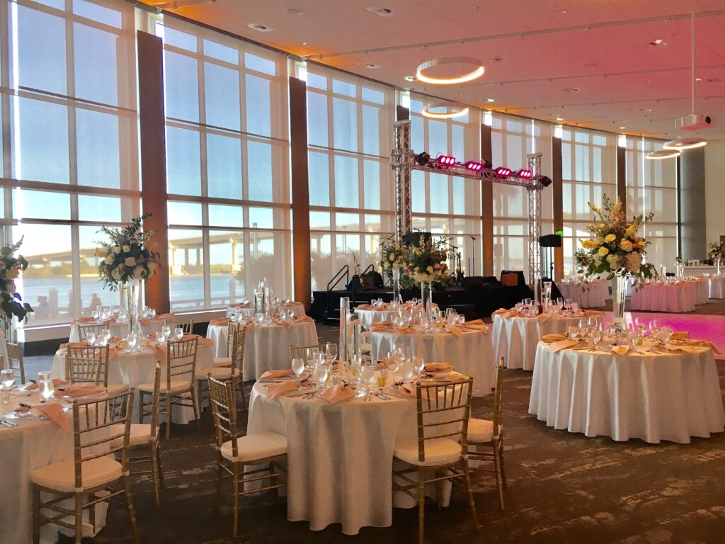 ballroom at brannon center florida set up for wedding reception with white tables and chairs and pink lighting in room with large windows overlooking the water